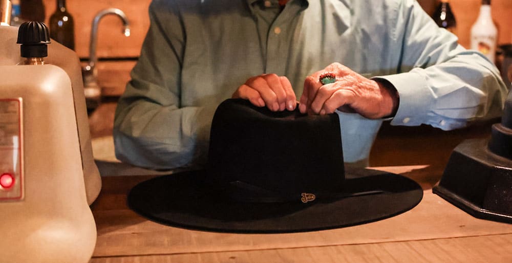 Un hombre con sus manos sobre un sombrero negro sobre el mostrador, dándole forma a un sombrero.